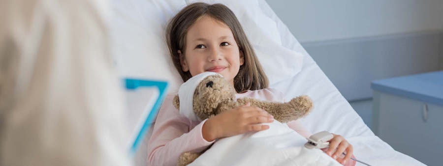 Mädchen liegt im Patientenbett, ihren Kuschelbären mit Kopfverband im Arm