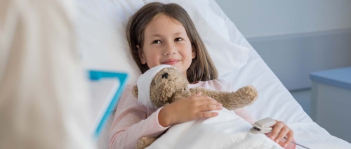 Mädchen liegt im Patientenbett, ihren Kuschelbären mit Kopfverband im Arm