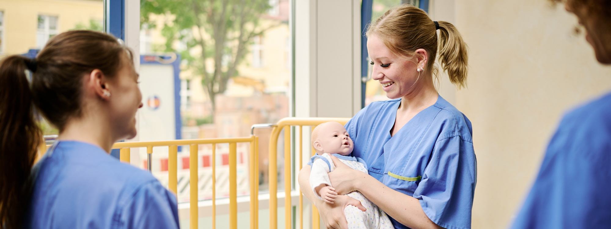 Hebammenstudentinnen sind zusammen mit Praxisanleiterin am Gitterbett, eine Studientin hält Babypuppe im Arm