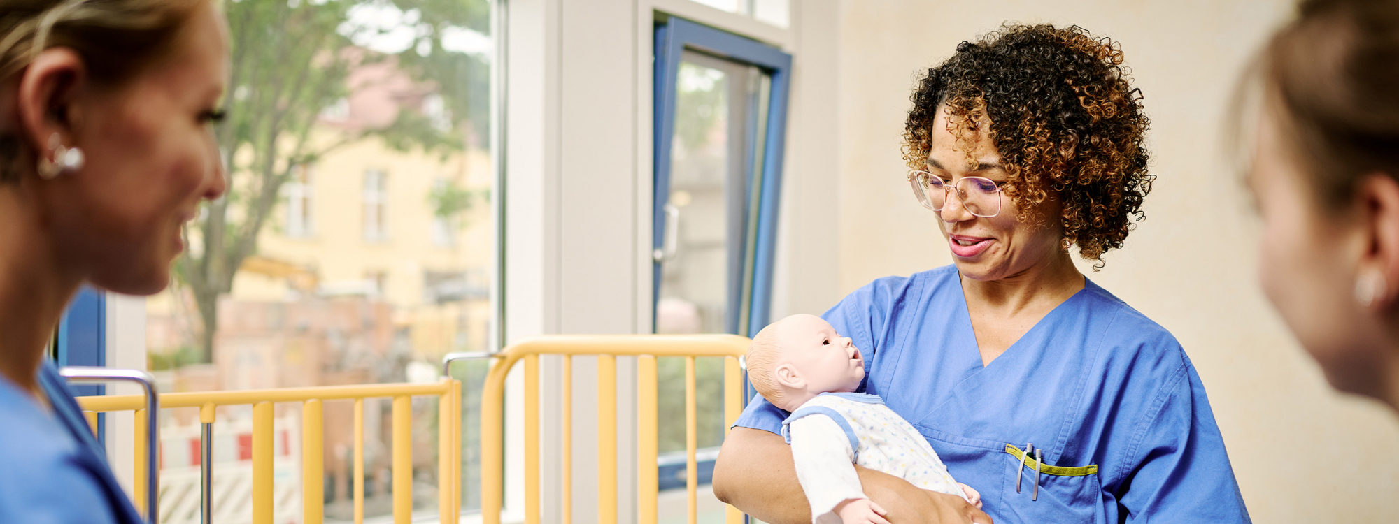 Praxisanleiterin hält Babypuppe im Arm, Hebammenstudentinnen schauen zu